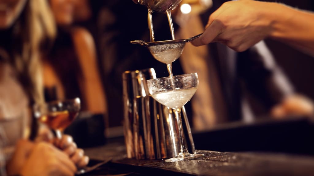 A bartender in California strains a cocktail into a coupe glass at the bar, with another cocktail and shakers visible in the background, perhaps contemplating the smooth transition of a liquor license transfer.